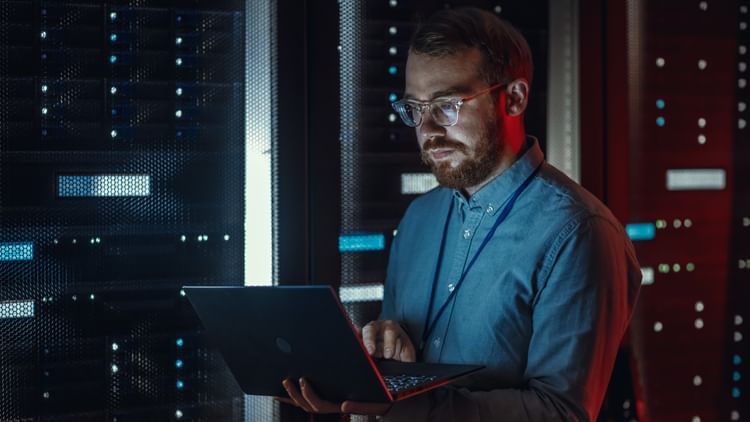 Bearded IT Specialist in Glasses is Working on Laptop in Data Center while Standing Near Server Rack